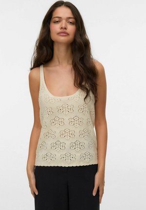 Kobieta o długich falowanych ciemnych włosach, ubrana w beżowy szydełkowany bezrękawnik i czarne spodnie, stojąca na tle jednolitego jasnego tła.