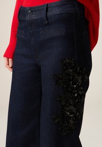 Donkerblauwe spijkerbroek met gedetailleerd zwart bloemrijk kraalwerk op de linker dij en een klassiek ontwerp met vijf zakken.