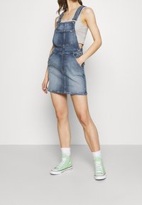 Kvinna som bär en ljusgrå linne med en blå denim overallkjol och ljusgröna högskor med vita strumpor.