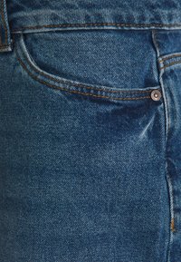 Modré džíny z denimu představují detailní pohled na texturu látky, šití a kovový knoflík poblíž kapsy.