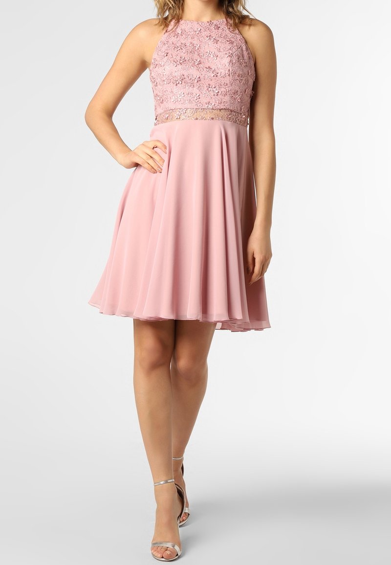 Femme portant une robe rose sans manches avec un corsage en dentelle et une jupe fluide, associée à des sandales argentées à talons hauts, debout devant un fond uni.