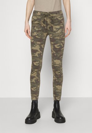 Personne portant un pantalon camouflage ajusté avec poches plaquées à l'avant, chevilles élastiquées, haut beige et bottines noires épaisses, debout devant un fond clair.