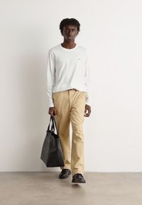 Cămașă albă cu mânecă lungă, pantaloni bej și pantofi negri. Poartă o geantă tote neagră. Design simplu, croială slim și aspect casual.