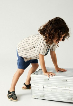 Bambino con capelli ricci che indossa una maglietta a righe e pantaloncini, che si sporge in avanti con le mani su una grande valigia argentata in un ambiente semplice.