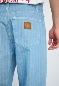 Lyseblå stripete jeans med et herringbone-mønster. Har en brun lærpatch med logo på baklommen og hvite sømmer.