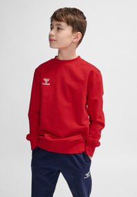 Felpa rossa a maniche lunghe realizzata in tessuto morbido, con scollo rotondo e un piccolo logo bianco sul petto. Abbinata a pantaloni blu navy.