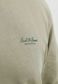 Gros plan sur un tissu beige avec un petit texte brodé en vert foncé « Jack & Jones Archive Selection » sur la partie supérieure arrière d'un vêtement.