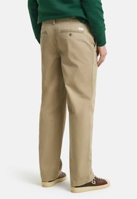 Beige cotton trousers with a relaxed fit, featuring back pockets with zippers, paired with dark brown sneakers and a green sweatshirt.