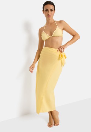 Woman wearing a yellow bikini top with ruffled edges and a matching long wrap skirt tied at the waist, standing barefoot against a white background.