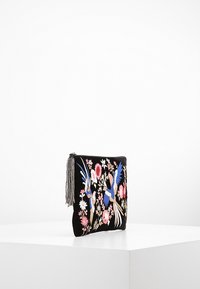 Czarna torebka kopertowa z kolorowymi haftowanymi ptakami i kwiatami, z metalowym suwadłem w postaci srebrnej frędzelki, wyeksponowana na białym piedestale.