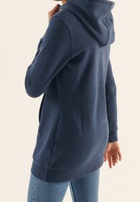 Tmavě modrá mikina s kapucí, se soft texturou, bočními kapsami a delším zadním lemem, v kombinaci se světle modrými džíny.