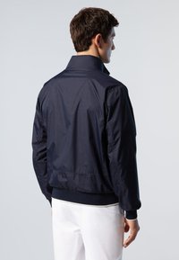 Giacca leggera blu navy con colletto alto, polsini elasticizzati e cinturino in vita. Texture liscia, design minimalista e senza modelli o accessori visibili.