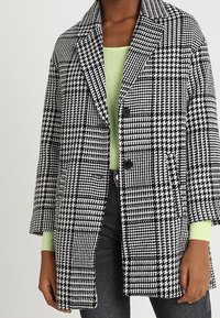 Cappotto a scacchi bianco e nero con colletto a notched, chiusura con due bottoni e vestibilità comoda, indossato sopra una maglietta verde chiaro, abbinato a pantaloni grigi.