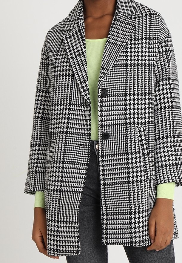 Cappotto a scacchi bianco e nero con colletto a notched, chiusura con due bottoni e vestibilità comoda, indossato sopra una maglietta verde chiaro, abbinato a pantaloni grigi.