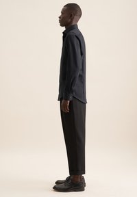 Maglietta nera a maniche lunghe con colletto strutturato, abbinata a pantaloni neri su misura e scarpe nere stringate, vista di lato.