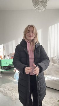 Νέα γυναίκα που διορθώνει ένα μαύρο μακρύ μπουφάν πάνω από ροζ hoodie, στέκεται σε φωτεινό σαλόνι με πράσινη ντουλάπα και πολυέλαιο.