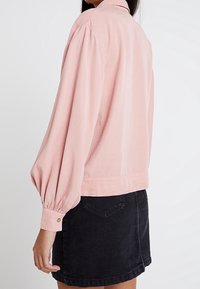 Giacca lunga rosa chiaro con colletto, maniche voluminose e polsini con bottoni, abbinata a una gonna di jeans nera.