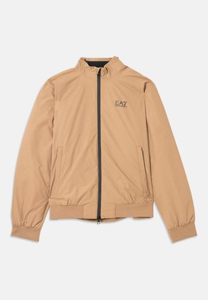 Lett beige jakke med høy krage, full glidelås foran og elastiske mansjetter. Har sidelommer og en liten svart logo på brystet.