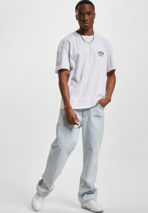 Hvit bomulls t-skjorte med en logo på venstre bryst, kombinert med lyseblå denimbukser. Modell som bruker hvite sneakers, står mot en ensfarget bakgrunn.