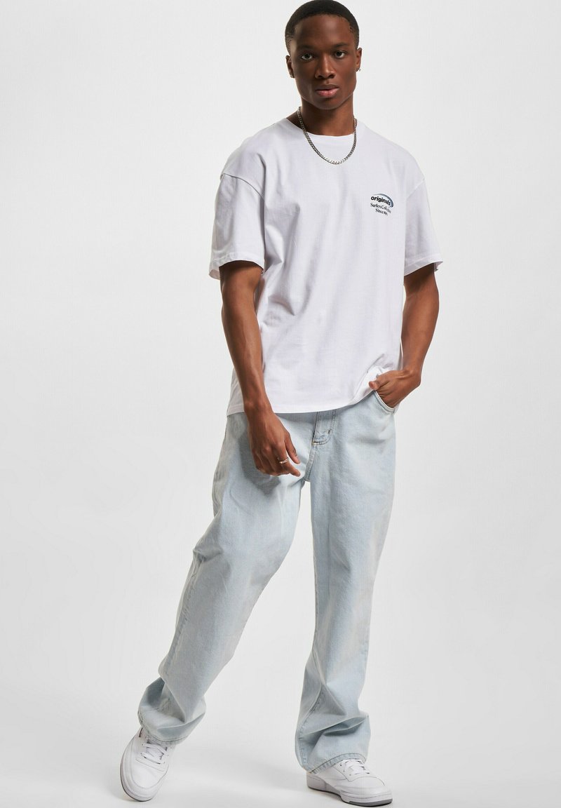 Hvit bomulls t-skjorte med en logo på venstre bryst, kombinert med lyseblå denimbukser. Modell som bruker hvite sneakers, står mot en ensfarget bakgrunn.