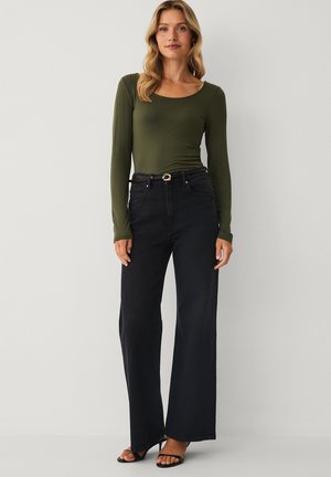 Woman wearing olive green long-sleeve top, high-waist black wide-leg jeans with a belt, and black open-toe heels standing against a plain background.