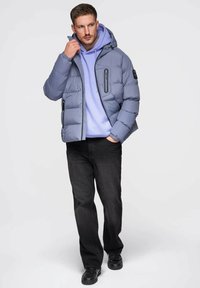 Szara kurtka puchowa z kapturem i czarnymi kieszeniami na zamek, noszona na jasnofioletowej bluzie z kapturem, w połączeniu z czarnymi spodniami o prostych nogawkach i czarnymi butami.