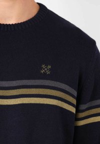 Pull en maille marine avec un col côtelé. Il présente des rayures horizontales grises et dorées, avec un petit design brodé sur la poitrine.