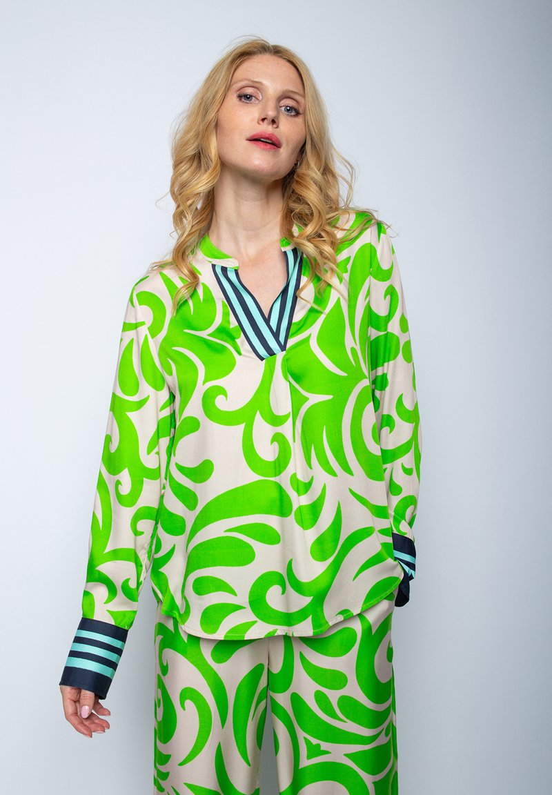 Zielona i beżowa bluzka w wzory z dekoltem w serek, z wirującymi wzorami i ciemnymi, prążkowanymi mankietami. Gładki materiał o luźnym kroju.