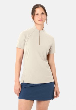 Junge Frau trägt ein beigefarbenes, kurzärmeliges Reißverschluss-Oberteil und einen blauen Rock, steht vor einem schlichten hellen Hintergrund.