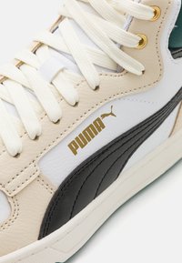 Gros plan sur une basket Puma blanche et beige avec une bande noire, des lacets crème et un logo Puma doré près des oeillets.