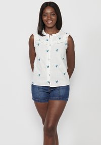Weißes ärmelloses Blusenshirt mit rüschigem Kragen, verziert mit blauer Blumenbestickung, kombiniert mit Jeansshorts. Glatte Textur, vorn mit Knopfverschluss.
