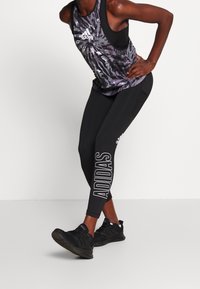 Czarne legginsy z napisem "ADIDAS" po boku, zestawione z wzorzystym topem bez rękawów w odcieniach szarości i czerni. Czarne buty sportowe.