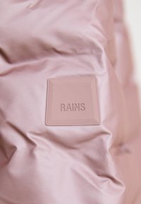 Šviesiai rožinė pūkuota medžiaga su lygiu paviršiumi, centre turi iškilų kvadratinį ženklą su įspaudu "RAINS".
