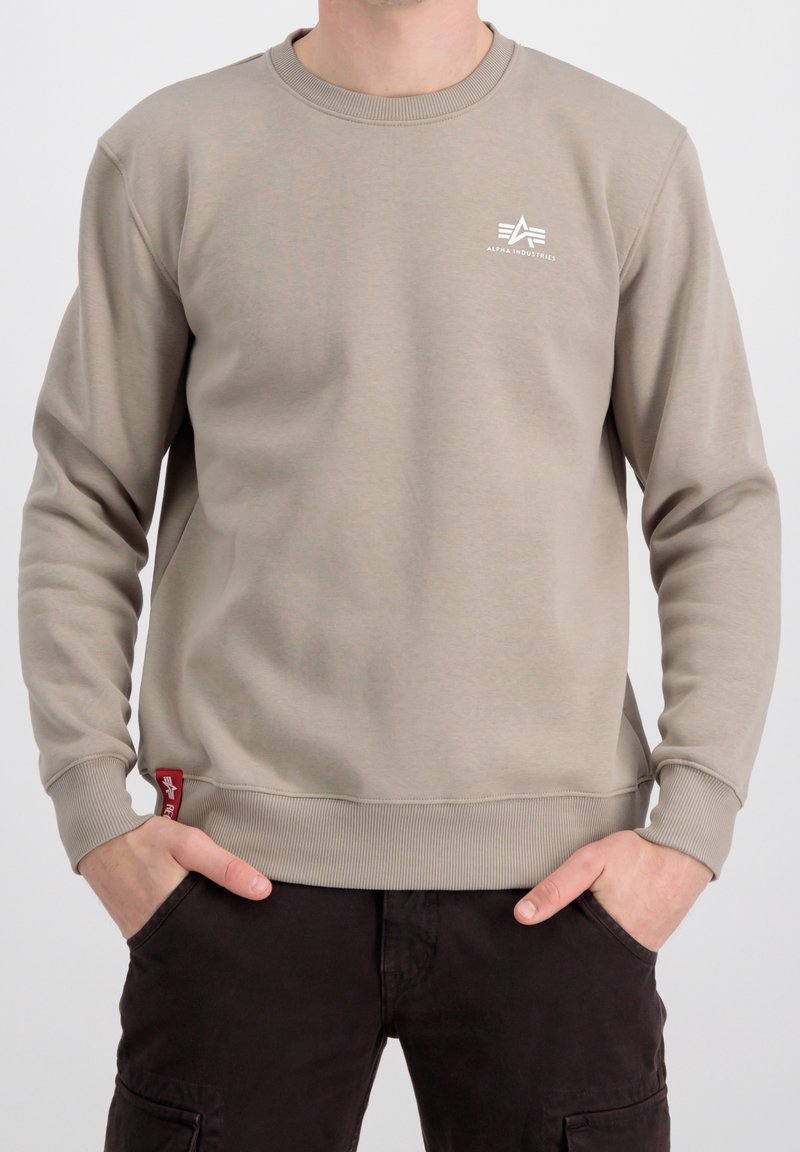 Homme portant un sweat-shirt beige à col rond avec un petit logo blanc Alpha Industries sur la poitrine et une étiquette de logo rouge près de l'ourlet, les mains dans les poches.