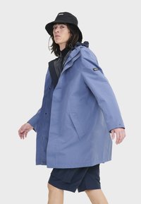 Parka impermeable azul con capucha, bolsillos frontales y marca en la manga. Combinada con pantalones cortos azul marino y un sombrero de cubo negro.