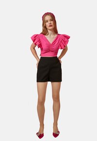 Rosa Cropped Top mit Rüschensleeves, kombiniert mit hoch taillierten schwarzen Shorts. Rosa verzierten Schuhe vervollständigen das Outfit.