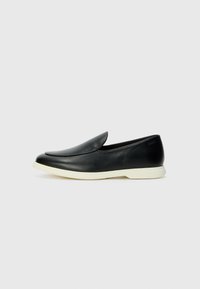 A fekete bőr slip-on cipők sima felülettel, lekerekített orral és fehér gumi talppal rendelkeznek, melyen diszkrét márkajelzés található a hátulján.
