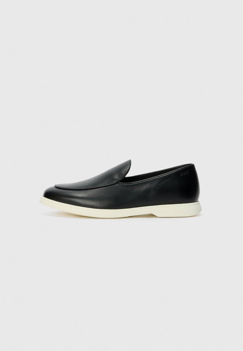 A fekete bőr slip-on cipők sima felülettel, lekerekített orral és fehér gumi talppal rendelkeznek, melyen diszkrét márkajelzés található a hátulján.