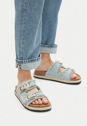 Jasnoniebieskie sandały na płaskiej podeszwie z denimu z postrzępionymi krawędziami, podwójnym zapięciem na klamry i korkową wkładką. Teksturowana gumowa podeszwa dla lepszej przyczepności.