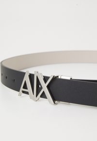 Sort læderbælte med sølv "AX" logo spænde, slankt design, glat overflade og flere huller til justering.