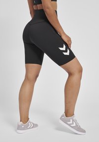 Svarta höga sportshorts i stretchigt tyg, med en vit chevronlogotyp på sidan och en slät, elegant yta.