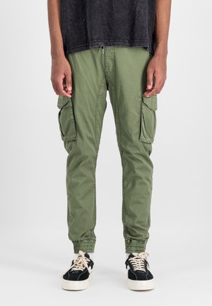 Green cargo pants with elasticized cuffs, two large side pockets, and a smooth cotton texture. Worn with black sneakers and a dark top.