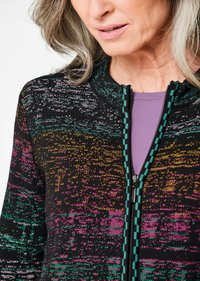 Zwarte zip-up jas met een multikleurig, textuurpatroon in tinten van roze, groen en goud; teal accenten langs de rits.