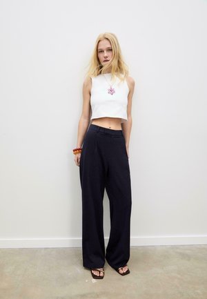 Weißes, gekürztes Top, marineblaue, weite Hosen und braune Sandalen. Eine rosa Halskette und ein Holzarmband setzen Akzente. Glatte Texturen, minimalistisches Design.