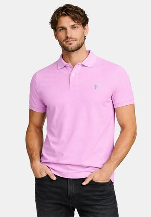 Mann mit Bart, der ein helllila kurzärmeliges Poloshirt und schwarze Jeans trägt, steht mit den Händen in den Taschen vor weißem Hintergrund.