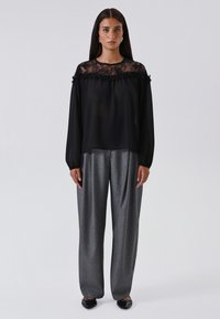 Una donna sta in piedi indossando una blusa di pizzo nero trasparente con motivi floreali e rouches, abbinata a pantaloni metallici argento dalla vestibilità ampia e ballerine nere.