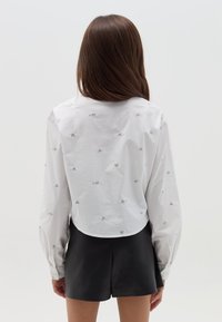 Camisa blanca recortada con bordado de corazón plateado, mangas largas, puños de botón y un dobladillo redondeado, combinada con pantalones cortos negros.