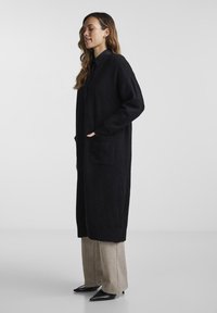 Svart lång cardigan i ett mjukt texturerat tyg, med två framfickor och knappknäppning, kombinerad med ljusbeige vida byxor.