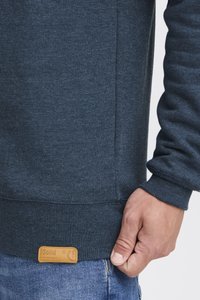 Sudadera azul marino con puños y dobladillo acanalados. Cuenta con una tela texturizada, un bolsillo lateral y una etiqueta de marca en tono marrón en el dobladillo inferior.