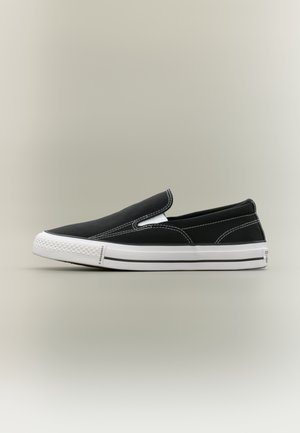 Baskets slip-on noires avec semelle en caoutchouc blanche et surpiqûres blanches contrastantes sur un fond neutre, vue de côté.